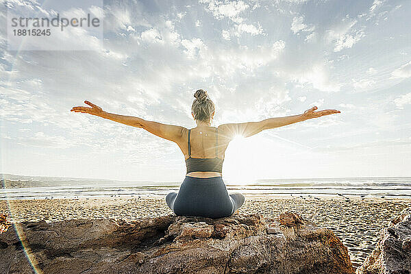 Frau praktiziert Yoga mit ausgestreckten Armen und sitzt auf einem Felsen am Strand vor dem Himmel