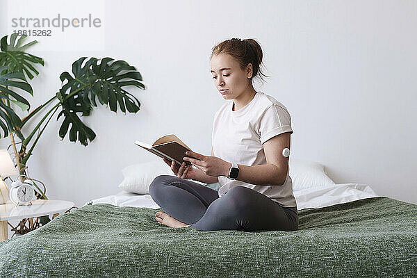 Frau mit Diabetes liest Buch und sitzt mit gekreuzten Beinen zu Hause im Bett