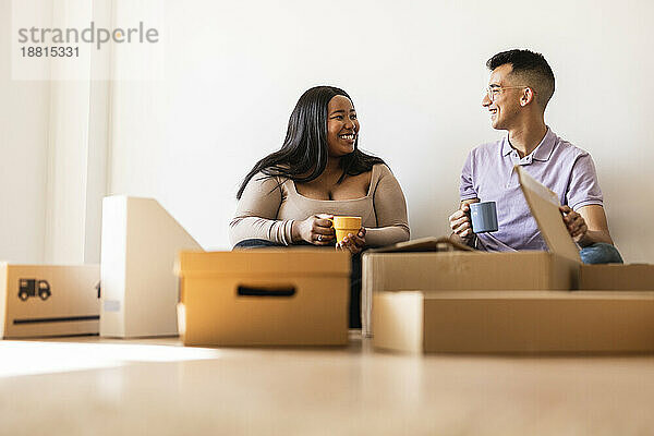 Lächelndes Paar mit Kaffeetasse sitzt neben Kartons im neuen Zuhause