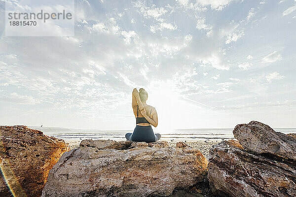 Frau praktiziert Yoga mit den Händen hinter dem Rücken und sitzt auf einem Felsen am Strand