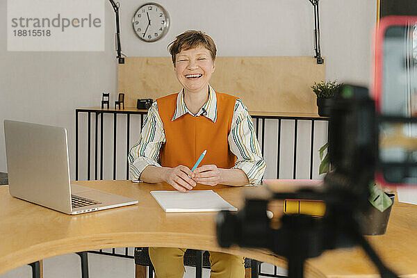 Lächelnde Frau sitzt im Klassenzimmer und blickt auf die Kamera  die den Online-Kurs aufzeichnet