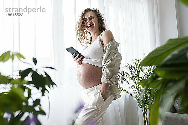 Happy pregnant woman with hand in pocket holding smart phone at home