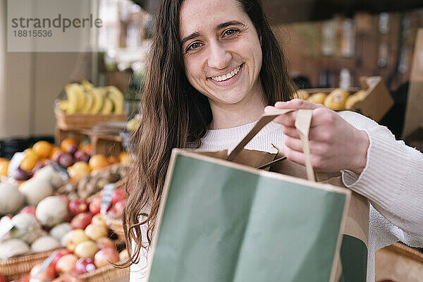 Glücklicher junger Lebensmittelhändler steht mit Papiertüte am Gemüsestand