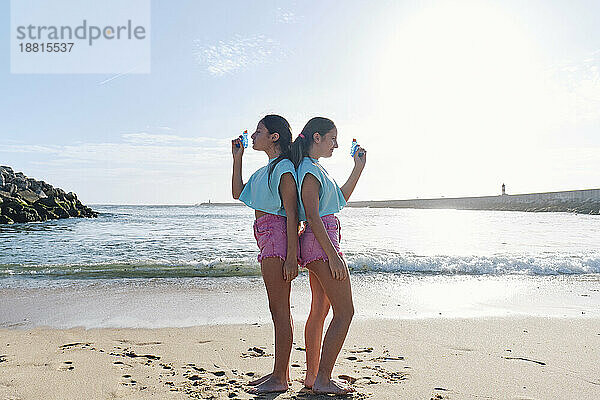 Zwillingsschwestern in passenden Outfits stehen Rücken an Rücken mit Spritzpistolen am Strand
