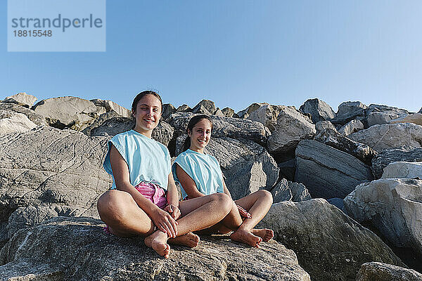 Smiling twin sisters wearing matching outfits sitting on rocks at beach