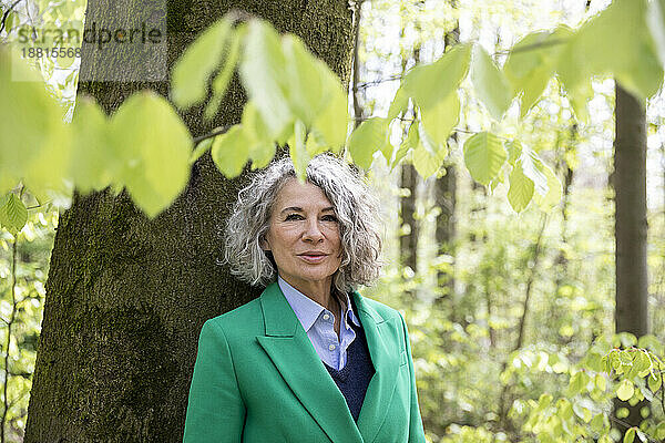 Ältere Frau lehnt an Baum im Wald und trägt einen grünen Mantel