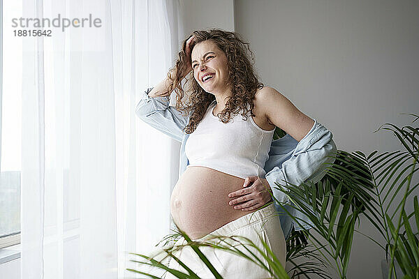 Happy pregnant woman with hand on hip standing near window at home