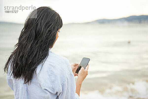 Frau mit langen Haaren benutzt Smartphone am Strand