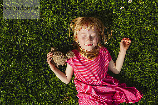 Lächelndes erdbeerblondes Mädchen mit geschlossenen Augen  das an einem sonnigen Tag im Gras liegt