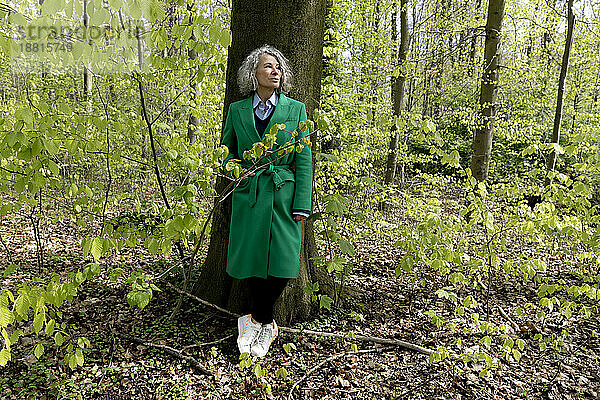 Ältere Frau lehnt an Baum im Wald und trägt grünen Mantel