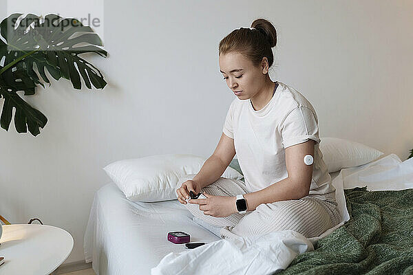 Young woman with diabetes checking glucose level sitting on bed at home