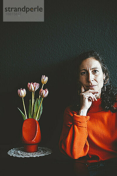 Frau mit Hand am Kinn lehnt an Tisch mit roter Blumenvase vor schwarzer Wand