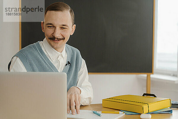 Lächelnder Mann sitzt im Klassenzimmer und schaut auf den Laptop