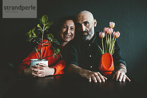 Älteres Paar mit Topfpflanze und Vase sitzt vor schwarzer Wand