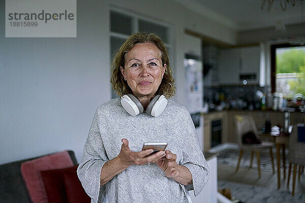 Lächelnde ältere Frau steht mit Smartphone zu Hause