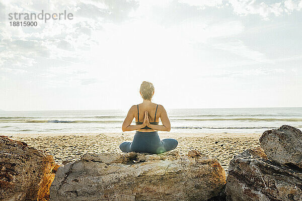 Frau praktiziert Yoga mit hinter dem Rücken verschränkten Händen und sitzt am Strand
