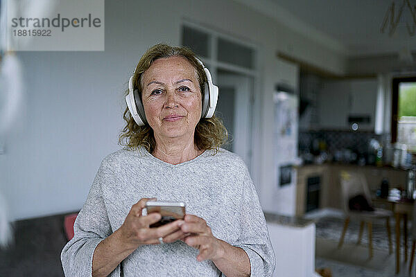 Ältere Frau mit kabellosen Kopfhörern steht zu Hause mit Smartphone