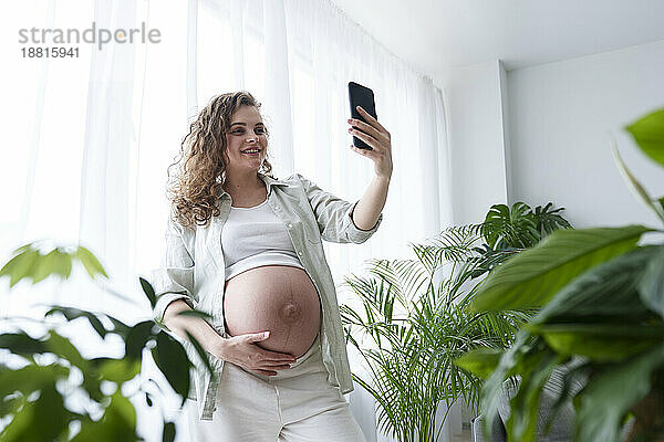 Lächelnde schwangere Frau macht ein Selfie mit dem Smartphone inmitten von Pflanzen zu Hause