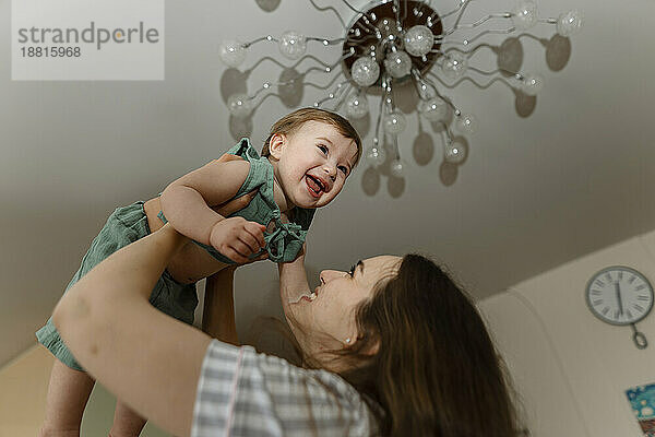 Happy mother lifting baby girl under ceiling at home