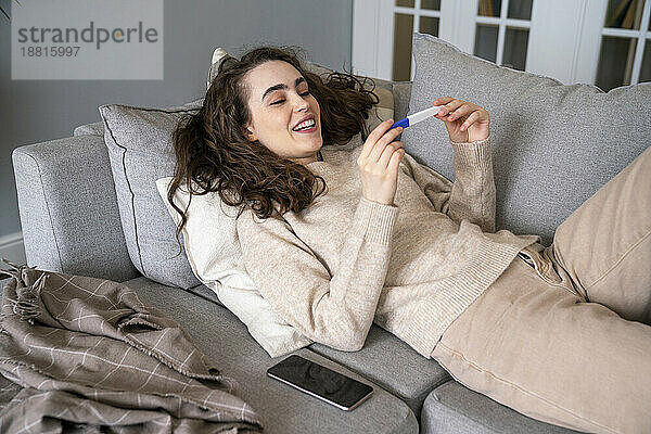 Glückliche junge Frau schaut auf das Schwangerschaftstest-Set und entspannt sich auf dem Sofa