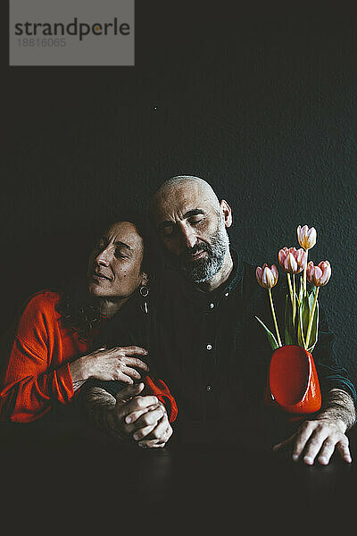 Romantisches Paar mit roter Blumenvase sitzt vor schwarzer Wand