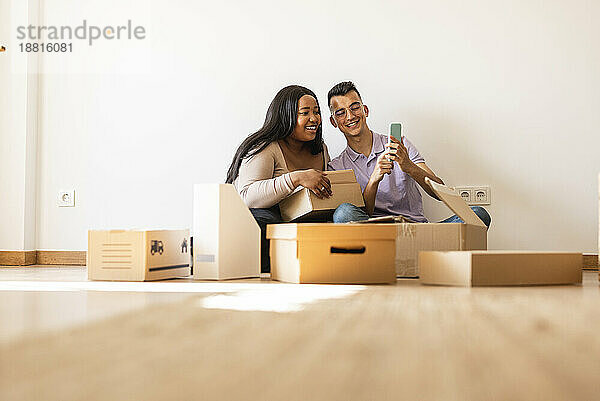 Glückliches Paar macht Selfie und sitzt neben Pappkartons im neuen Zuhause