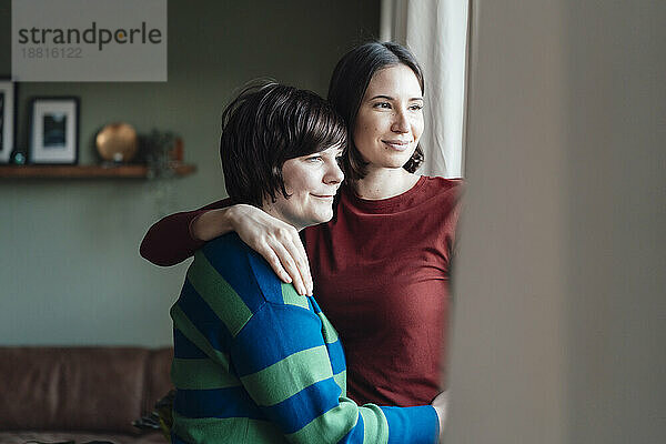 Smiling woman with arm around lesbian friend looking out of window at home