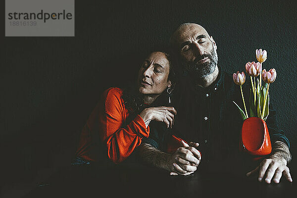 Couple with eyes closed sitting with red flower vase together in front of black wall