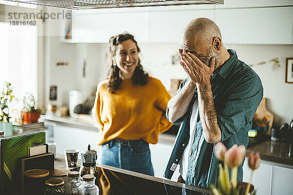 Mann bedeckt Gesicht mit Händen und steht in der Küche neben einer lachenden Frau