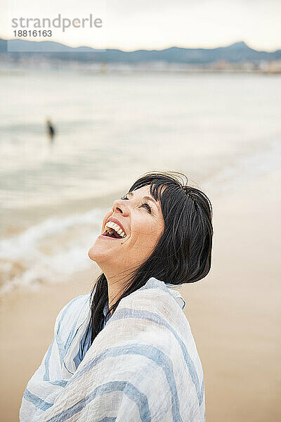 Glückliche Frau  in einen Schal gehüllt  lacht am Strand