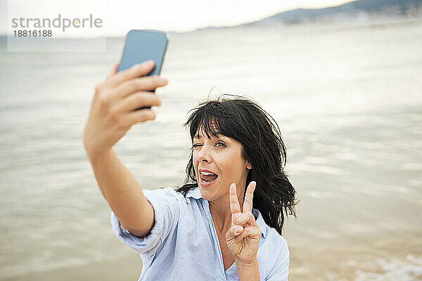 Frau macht ein Friedenszeichen und macht ein Selfie mit ihrem Smartphone am Strand