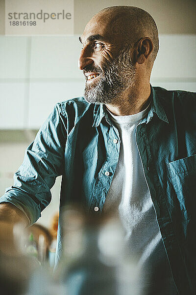 Lächelnder reifer Mann mit rasiertem Kopf und ergrauendem Bart sitzt in der Küche