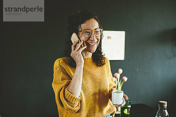 Lächelnde Frau  die zu Hause mit einem Smartphone spricht und eine Tasse hält