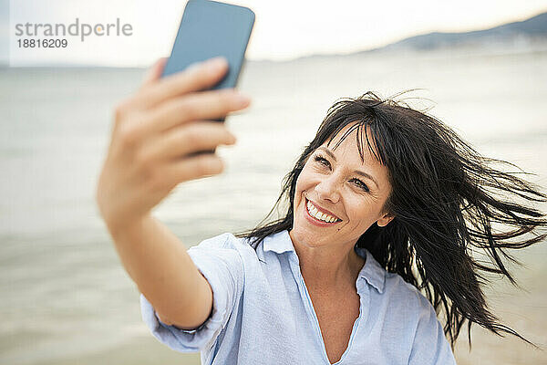 Glückliche Frau mit zerzausten Haaren  die am Strand ein Selfie mit dem Smartphone macht