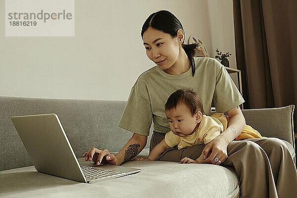 Freiberufliche Mutter benutzt Laptop und sitzt mit ihrer Tochter zu Hause auf dem Sofa