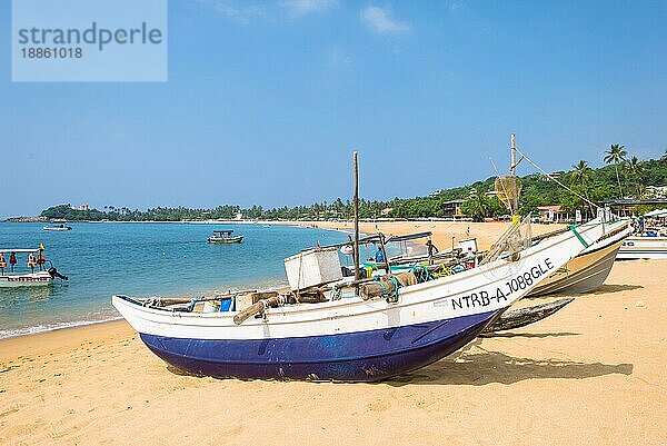 Am Strand eines der wichtigsten Touristenzentren im Südwesten Sri Lankas. Touristen nehmen ein Sonnenbad und treiben Wassersport. Auslegerboote und traditionelle Fischerboote am Strand und vor Anker