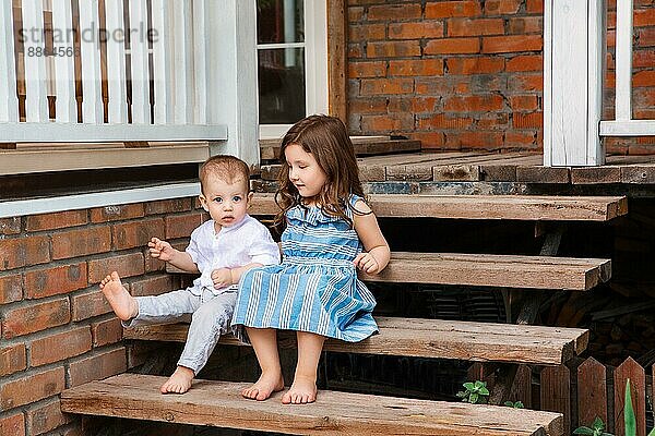 Niedliche kleine Bruder und Schwester sitzen auf Holztreppe in der Nähe Veranda des Landhauses und entspannen in der ländlichen Gegend während der Sommerferien