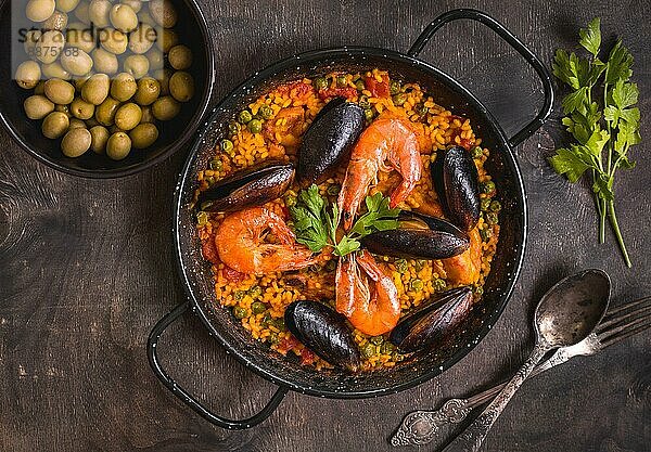 Paella in schwarzer Pfanne mit Reis  Krabben  Muscheln  Tintenfisch und Fleisch  Schüssel mit Oliven und Vintage-Besteck. Paella mit Meeresfrüchten  traditionelles spanisches Gericht. Paella auf rustikalem Tisch. Selektiver Fokus. Ansicht von oben