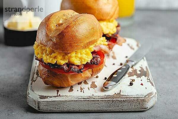 Frühstückssandwiches mit Rührei  Speck  Käse  Tomaten auf weißem Holzbrett  Glas mit frischem Orangensaft  weißer Hintergrund. Frühstück machen Konzept  selektiver Fokus