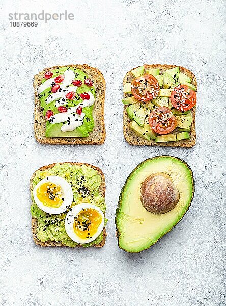 Avocado-Toasts mit Ei  Tomaten  Gewürzen und einer halben ganzen Avocado auf weißem Steinhintergrund. Gesunde Frühstücks-Avocado-Sandwiches mit verschiedenen Belägen  Draufsicht  Nahaufnahme