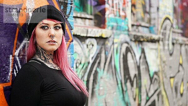 Junge urbane Frau mit rosa Haaren  Piercings und Tattoos lehnt an einer grafitti Wand - echte Menschen alternativer Lebensstil