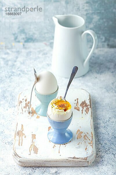 Frische weich gekochte Eier in Ständen  Löffel  weiße Holzkochplatte  weißer Beton rustikalen Hintergrund. Weiche Eier für gesundes Frühstück. Glas mit Milch. Selektiver Fokus. Eiweiß-Fitness-Frühstück