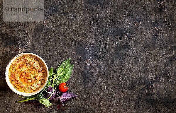 Hummus auf einem Teller mit Kirschtomaten und Kräutern auf einem dunklen Holzhintergrund. Gericht aus dem Nahen Osten. Platz für Text