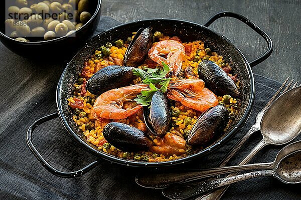 Paella in schwarzer Pfanne mit Reis  Krabben  Muscheln  Tintenfisch und Fleisch  Schüssel mit Oliven und Vintage Besteck. Paella mit Meeresfrüchten  traditionelles spanisches Gericht. Paella auf einem rustikalen schwarzen Holztisch. Selektiver Fokus