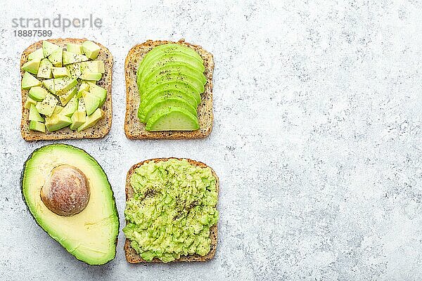 Verschiedene Arten von Avocado-Toasts und eine halbe ganze Avocado auf weißem Stein-Hintergrund  Platz für Text. Wie man ein leckeres gesundes Frühstücks-Avocado-Sandwich zubereitet und serviert  Draufsicht  Nahaufnahme