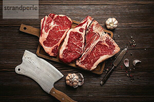 Set von drei verschiedenen Arten von rohem Fleisch Steaks: Ribeye  T-Bone  Cowboy auf Schneidebrett mit Messer und Gewürzen  Holzhintergrund. Gealtertes Steaksortiment  Metzgerei/Restaurantkonzept  Draufsicht  Nahaufnahme