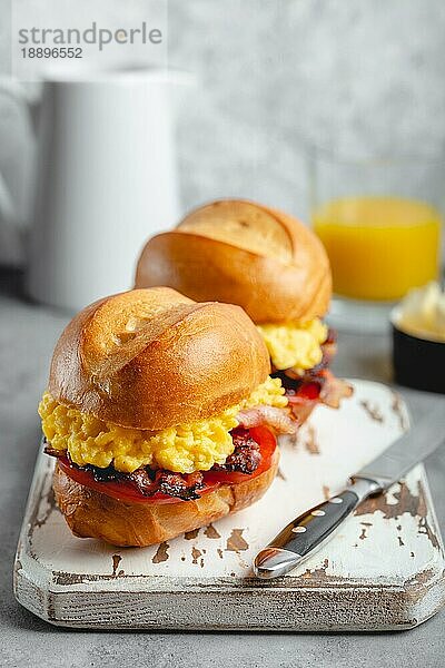 Frühstückssandwiches mit Rührei  Speck  Käse  Tomaten auf weißem Holzbrett  Glas mit frischem Orangensaft  weißer Hintergrund. Frühstück machen Konzept  selektiver Fokus