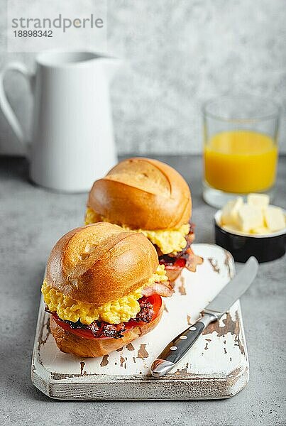 Frühstückssandwiches mit Rührei  Speck  Käse  Tomaten auf weißem Holzbrett  Glas mit frischem Orangensaft  weißer Hintergrund. Frühstück machen Konzept  selektiver Fokus