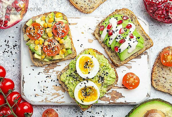 Avocado-Toasts mit Ei  Tomaten  Gewürze auf weißem Holz rustikalen Schneidebrett  Stein Hintergrund. Zutaten für gesunde Frühstücks-Avocado-Sandwiches mit verschiedenen Belägen  Draufsicht  Nahaufnahme