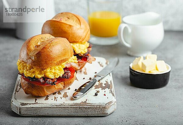 Frühstückssandwiches mit Rührei  Speck  Käse  Tomaten auf weißem Holzbrett  Glas mit frischem Orangensaft  weißer Hintergrund. Frühstück machen Konzept  selektiver Fokus